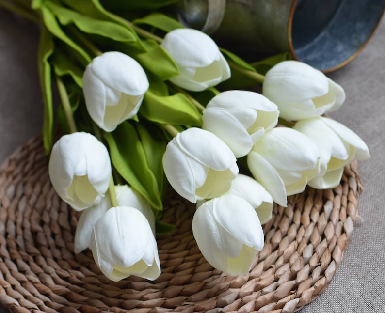 May include: A bouquet of white artificial tulips arranged on a woven wicker placemat. The tulips are made of a soft, realistic material and have a natural, elegant look.