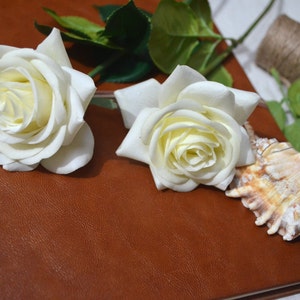 May include: Two white roses and a seashell on a brown leather journal.