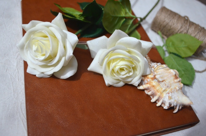 May include: Two white roses and a seashell on a brown leather journal.