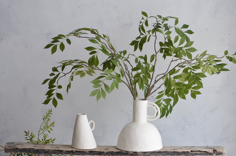 May include: Two white ceramic vases with a leafy green plant in the larger vase. The smaller vase is cone-shaped with a handle. The larger vase has a wide base and a handle on the side.