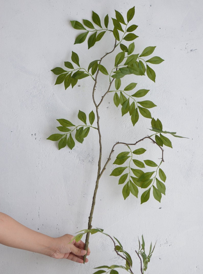 May include: A single artificial branch with green leaves, the branch is brown and has a natural look. The branch is held in a hand against a white background.