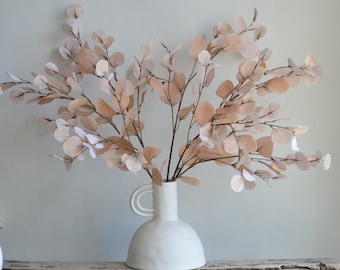 May include: A bouquet of dried eucalyptus branches with light brown leaves in a white ceramic vase with two handles. The vase has a rounded base and a wide opening.