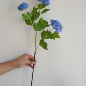 May include: A single stem of artificial blue hydrangea flowers with green leaves. The stem is brown and has a natural wood look.