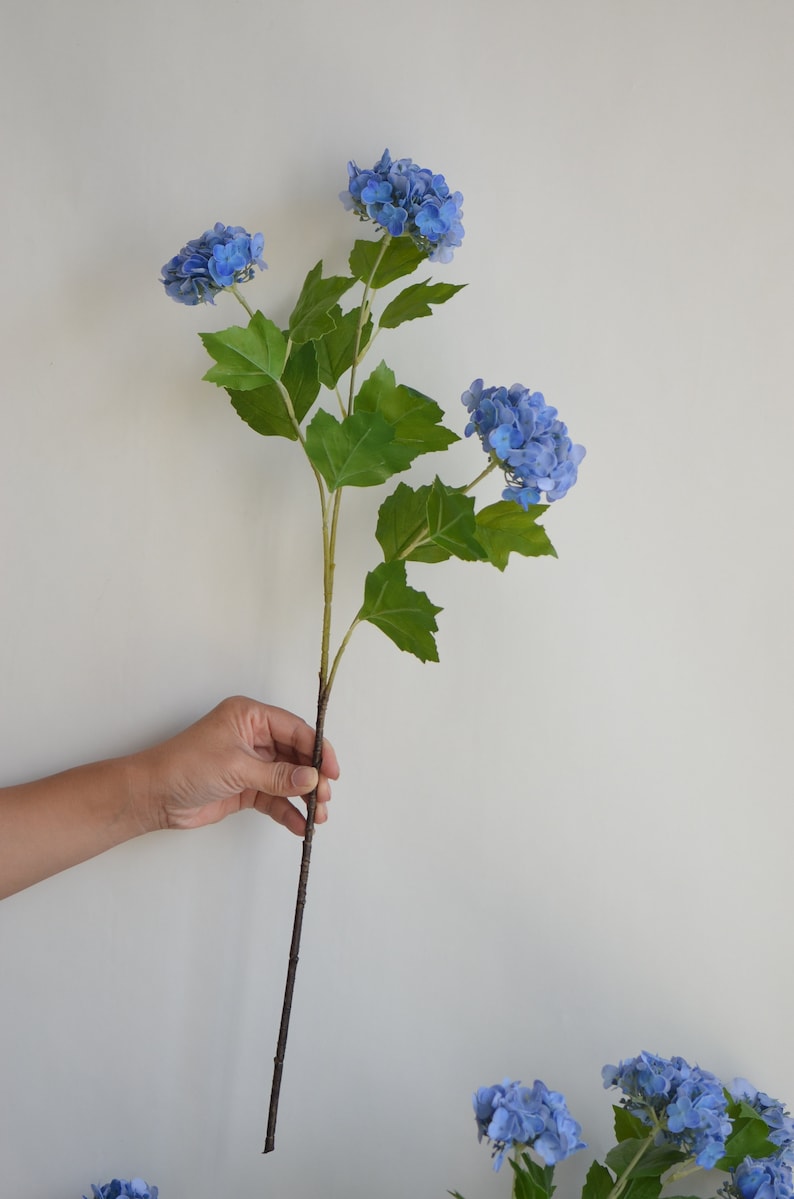 May include: A single stem of artificial blue hydrangea flowers with green leaves. The stem is brown and has a natural wood look.