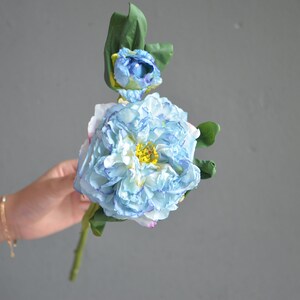 May include: A single stem artificial flower with a large blue and white flower and a smaller blue flower bud. The flower has a yellow center.