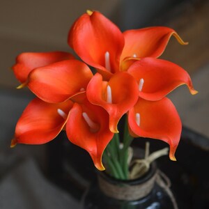 May include: A bouquet of orange calla lilies in a vase. The lilies are arranged in a cluster, with the stems visible. The vase is brown and has a rustic look.