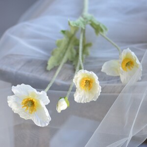 Puede incluir: Un ramo de flores de amapola de seda blanca con centros amarillos. Las flores est&aacute;n dispuestas sobre un fondo de tela blanca.