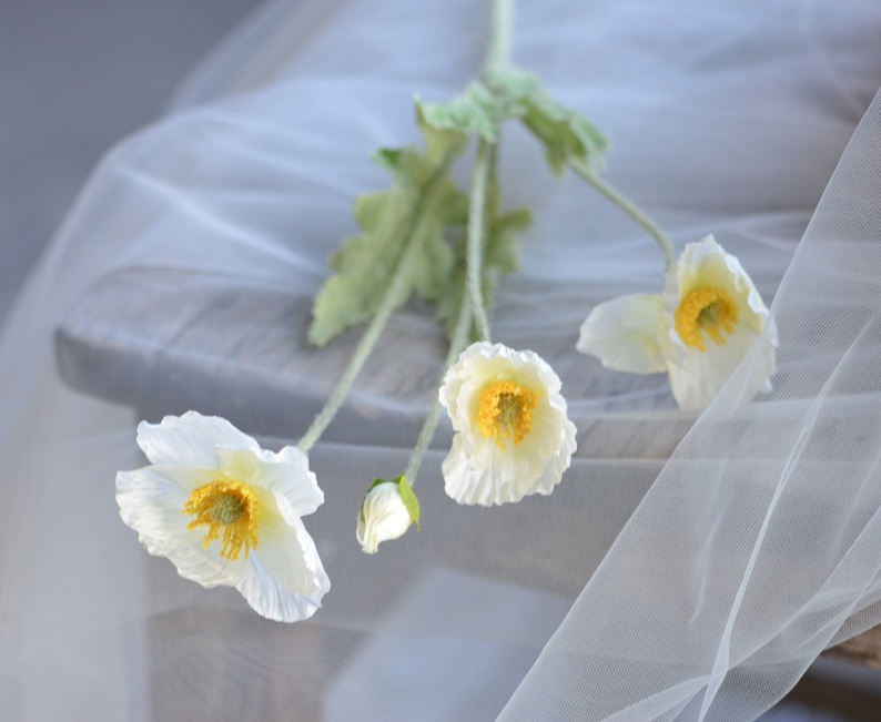 Puede incluir: Un ramo de flores de amapola de seda blanca con centros amarillos. Las flores est&aacute;n dispuestas sobre un fondo de tela blanca.