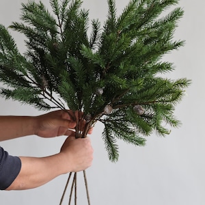 May include: A bundle of faux evergreen branches with realistic green needles and small pinecones. The stems are brown and held together, creating a natural, decorative arrangement. Perfect for holiday or seasonal decor.
