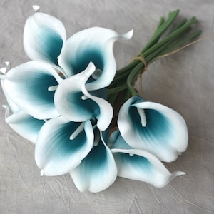 May include: A bouquet of artificial calla lilies. The flowers have white petals with a teal gradient towards the center. The stems are green and tied together with twine. The flowers are arranged in a bundle.