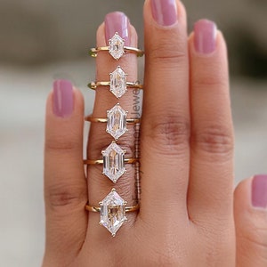 May include: A set of seven gold rings with different sized emerald cut diamonds. The rings are arranged in order of size, with the smallest ring on the left and the largest ring on the right.