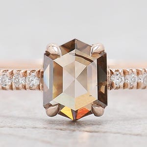 Op de afbeelding: Een ros&eacute;gouden verlovingsring met een zeshoekige, lichtbruine diamant. De band is bezet met kleine, heldere accentstenen. De ring wordt gepresenteerd tegen een zachte, neutrale achtergrond.
