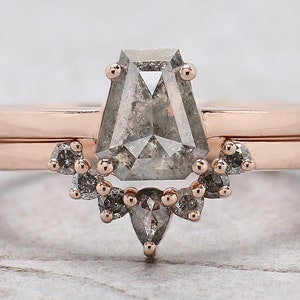 May include: A rose gold engagement ring set featuring a geometric, gray diamond center stone. The ring is paired with a simple band and a curved band with smaller gray stones, creating a unique bridal set.