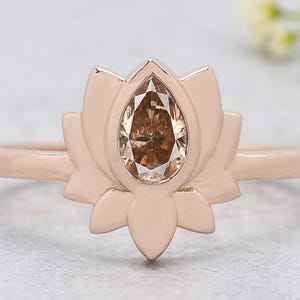 Op de afbeelding: Een roségouden ring met een lotusbloemdesign. De ring heeft een peervormige, lichtbruine edelsteen in het midden. De lotusblaadjes zijn gedetailleerd en omringen de edelsteen. De band is een eenvoudige, dunne band.
