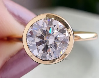 Op de afbeelding: Een gouden verlovingsring met een grote, ronde, heldere diamant. De ring wordt door een persoon vastgehouden. De band van de ring is dun en goudkleurig. De diamant is het middelpunt van de ring. Klassiek ontwerp.