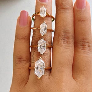May include: Four gold rings with elongated, clear, gemstone centerpieces are displayed on a hand with pink nail polish. The rings are of varying sizes, showcasing the different dimensions of the gemstones.
