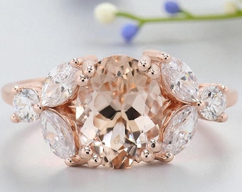 May include: Rose gold ring with a large, oval peach-coloured gemstone, framed by smaller, pear-shaped white stones. The ring's band is rose gold, and the setting has a floral design. The ring is set against a soft background.