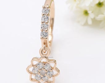 Può includere: Un singolo orecchino in oro rosa con un ciondolo a forma di fiore pendente. L'orecchino presenta una fila di piccole pietre preziose trasparenti lungo il cerchio. Il ciondolo a forma di fiore è anch'esso adornato con piccole pietre preziose trasparenti.