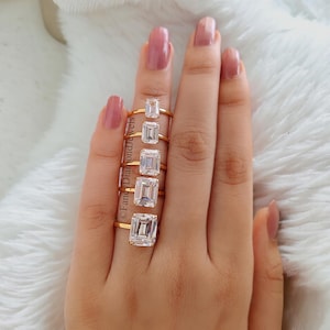 Puede incluir: Un conjunto de seis anillos de oro con piedras preciosas transparentes de talla esmeralda. Los anillos están apilados en un dedo, mostrando los diferentes tamaños de las piedras preciosas.