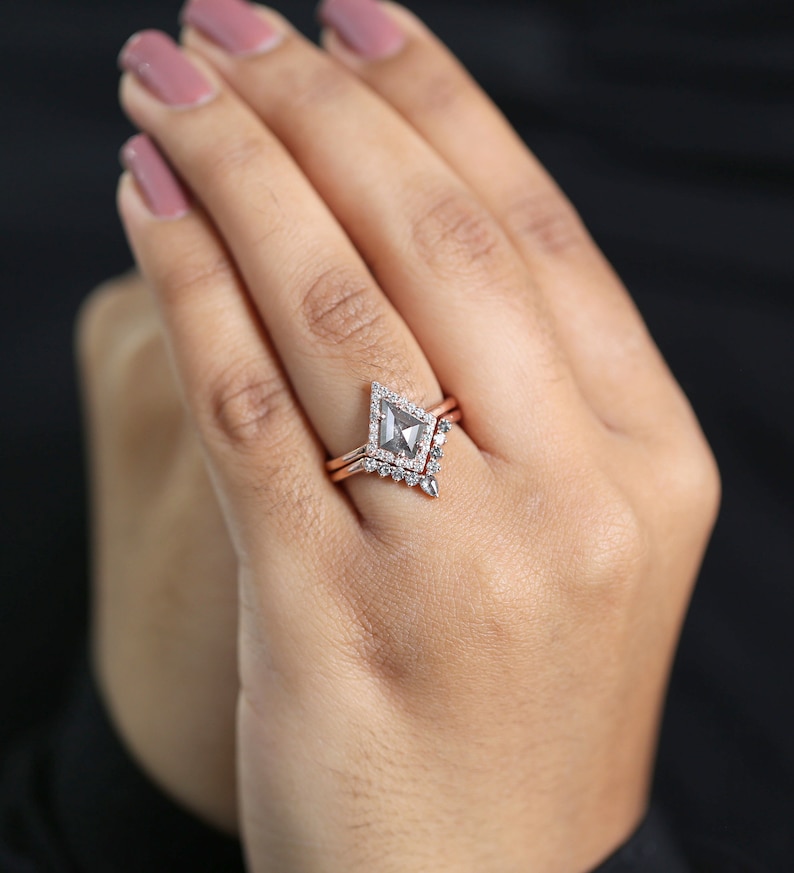 May include: A rose gold engagement ring set with a gray kite-shaped center stone and a diamond halo. The set includes a matching band with small diamonds. The ring is shown on a hand with pink nail polish.