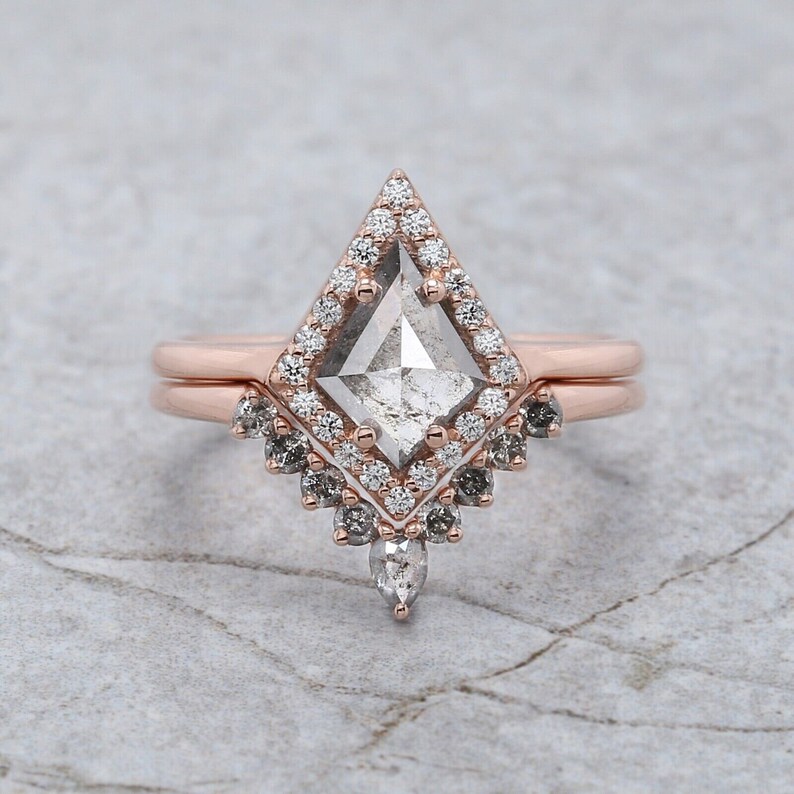 May include: Rose gold engagement ring set featuring a kite-shaped center stone surrounded by a halo of small diamonds. The band includes a matching contoured band with additional accent stones, creating a unique and elegant design. The ring is set on a light gray surface.