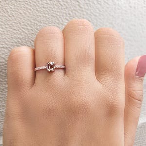 Op de afbeelding: Een ros&eacute;gouden ring met een rechthoekige, lichtbruine edelsteen als middelpunt. De band is versierd met kleine, heldere accentstenen. De ring wordt getoond op een hand met lichtroze nagellak.