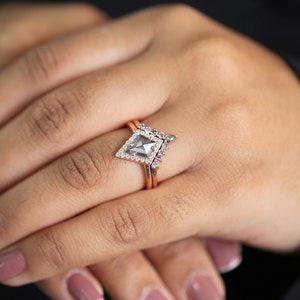 May include: A rose gold ring set featuring a geometric, kite-shaped center stone with a halo of small, clear stones. The main ring has a matching band. The rings are on a person's finger.