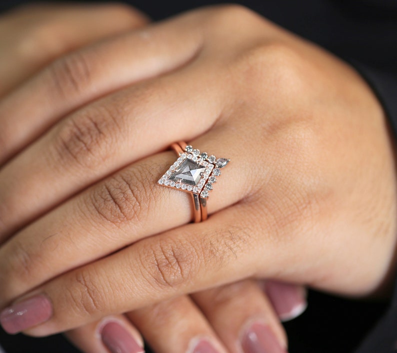 May include: A rose gold ring set featuring a geometric, kite-shaped center stone with a halo of small, clear stones. The main ring has a matching band. The rings are on a person's finger.