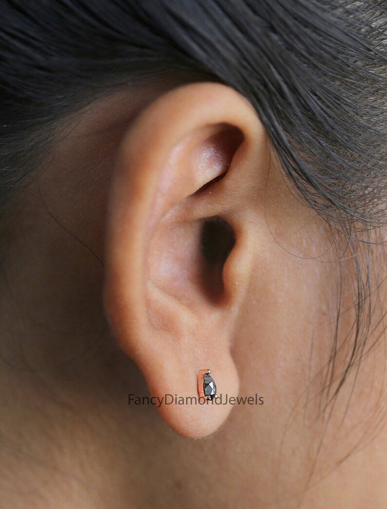 Puede incluir: Primer plano de una oreja con un peque&ntilde;o pendiente de diamante negro. El pendiente est&aacute; engastado en un engaste de oro rosa. El texto "FancyDiamondJewels" es visible debajo del pendiente.