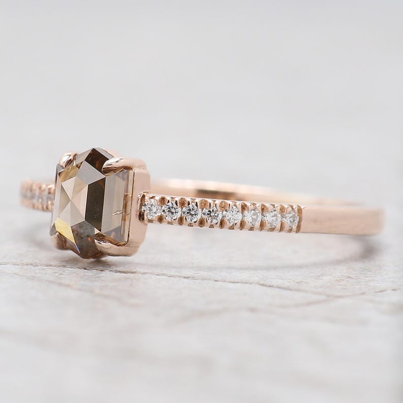 Op de afbeelding: Rosegouden verlovingsring met een rechthoekige, lichtbruine edelsteen. De band is versierd met kleine, heldere edelstenen. De ring staat tegen een zachte, vage achtergrond, die het ontwerp benadrukt.