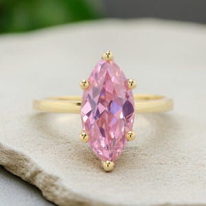 Puede incluir: Un anillo de oro con una piedra preciosa rosa en forma de marquesa. El anillo est&aacute; engastado con peque&ntilde;os detalles dorados alrededor de la piedra preciosa.