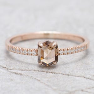 Op de afbeelding: Een ros&eacute;gouden ring met een zeshoekige, lichtbruine edelsteen als middelpunt. De band is versierd met kleine, heldere edelstenen. De ring is geplaatst tegen een lichtgrijze, getextureerde achtergrond.