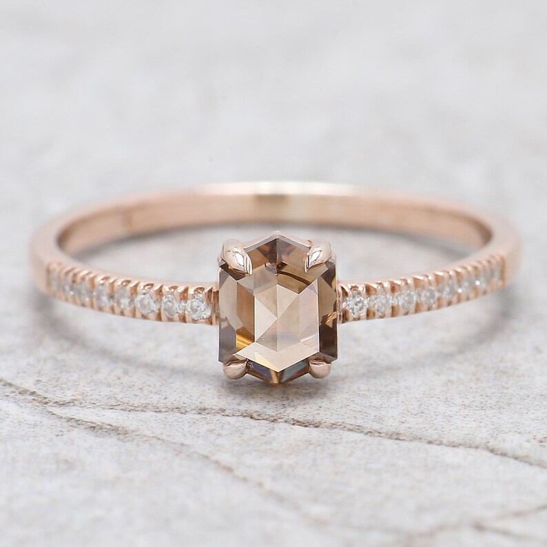 Op de afbeelding: Een ros&eacute;gouden ring met een zeshoekige, lichtbruine edelsteen als middelpunt. De band is versierd met kleine, heldere edelstenen. De ring is geplaatst tegen een lichtgrijze, getextureerde achtergrond.