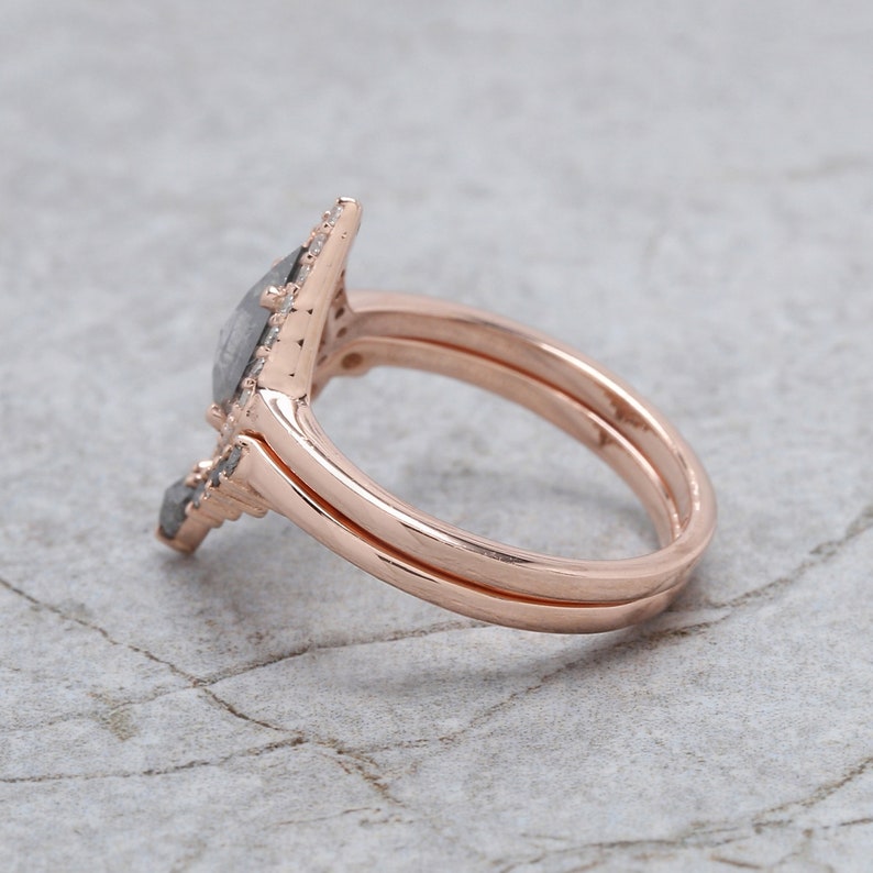 May include: Rose gold engagement ring set with a pear-shaped, gray diamond center stone and a matching band. The ring features a unique, angular design with smaller accent stones, set against a textured background.