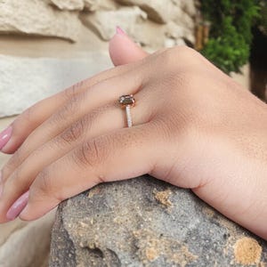 Op de afbeelding: Een ros&eacute;gouden ring met een rechthoekige, olijfgroene edelsteen. De band is bezet met kleine, heldere stenen. De ring zit om een vinger en rust op een stenen oppervlak.