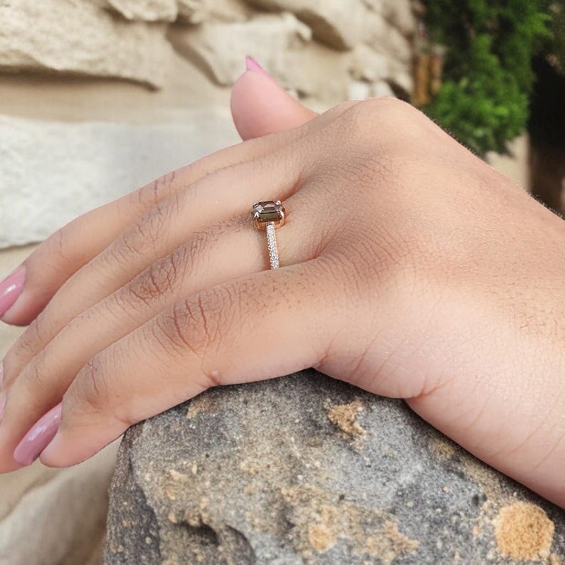 Op de afbeelding: Een ros&eacute;gouden ring met een rechthoekige, olijfgroene edelsteen. De band is bezet met kleine, heldere stenen. De ring zit om een vinger en rust op een stenen oppervlak.
