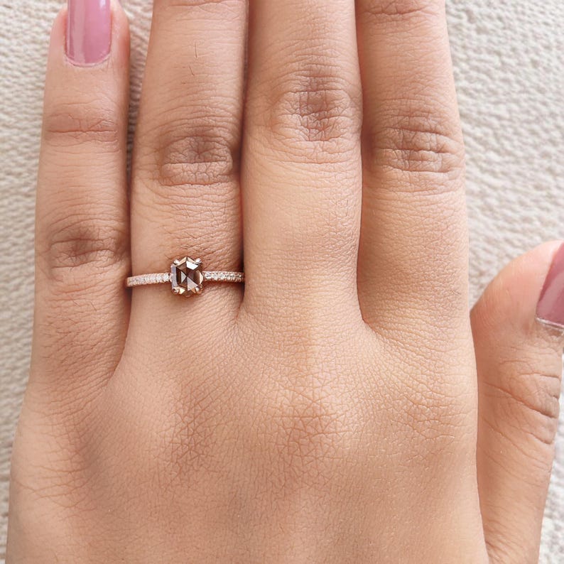 Op de afbeelding: Rosegouden ring met een vierkant geslepen, lichtbruine edelsteen. De band is versierd met kleine, heldere edelstenen. De ring wordt getoond op een hand met roze nagellak.
