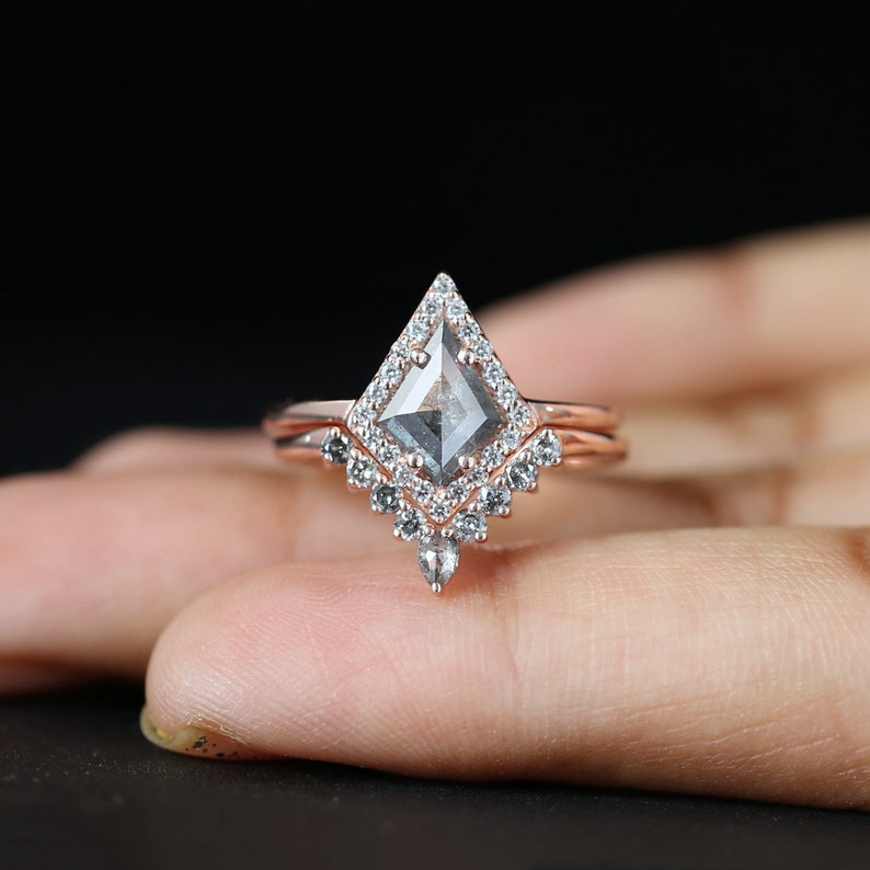 May include: A rose gold engagement ring set with a kite-shaped, gray-toned center stone. The ring is surrounded by a halo of small, clear accent stones. A matching band sits below the main ring, creating a layered effect. The background is black.