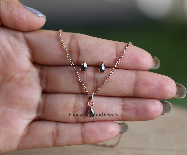 Puede incluir: Un collar con colgante de diamante negro y pendientes a juego en oro rosa. El colgante tiene forma de pera y los pendientes son peque&ntilde;os y redondos. El collar es delicado y tiene una cadena sencilla.