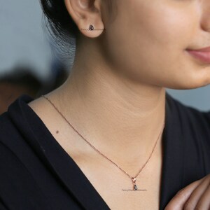 Puede incluir: Un delicado collar de oro rosa con un peque&ntilde;o colgante en forma de l&aacute;grima con una piedra preciosa negra. El collar lo lleva una persona con una camisa negra.