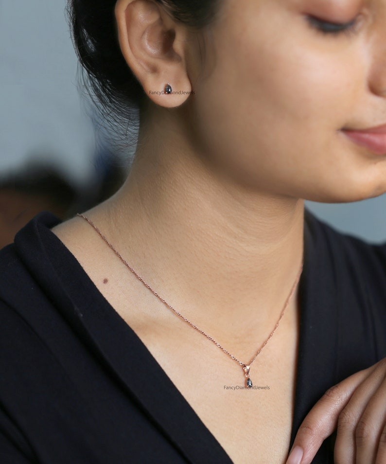 Puede incluir: Un delicado collar de oro rosa con un peque&ntilde;o colgante en forma de l&aacute;grima con una piedra preciosa negra. El collar lo lleva una persona con una camisa negra.