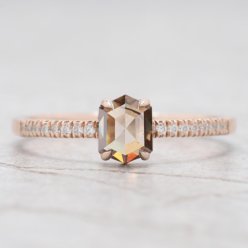 Op de afbeelding: Een ros&eacute;gouden verlovingsring met een zeshoekige, lichtbruine diamant. De band is bezet met kleine, heldere accentstenen. De ring wordt gepresenteerd tegen een zachte, neutrale achtergrond.