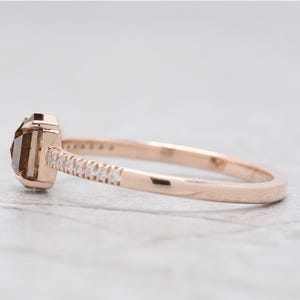 Op de afbeelding: Ros&eacute;gouden ring met een rechthoekige, bruine edelsteen. De band is versierd met kleine, heldere edelstenen. De ring wordt gepresenteerd tegen een neutrale achtergrond, wat het elegante ontwerp benadrukt. Een sieraad.