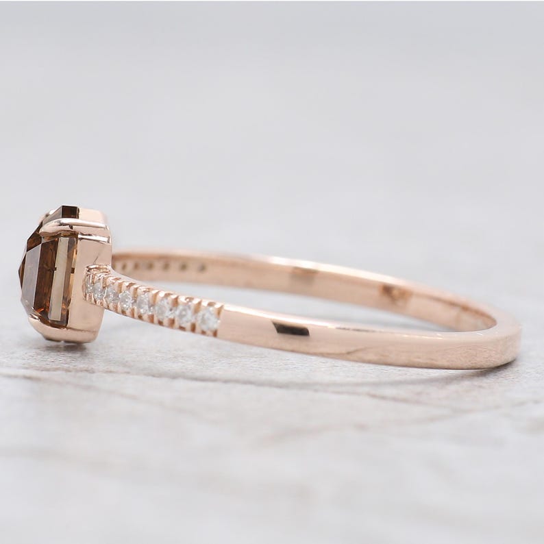 Op de afbeelding: Ros&eacute;gouden ring met een rechthoekige, bruine edelsteen. De band is versierd met kleine, heldere edelstenen. De ring wordt gepresenteerd tegen een neutrale achtergrond, wat het elegante ontwerp benadrukt. Een sieraad.