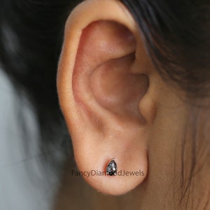 Puede incluir: Primer plano de una oreja con un peque&ntilde;o pendiente de diamante negro en forma de pera. El pendiente est&aacute; engastado en un engaste de plata u oro blanco.