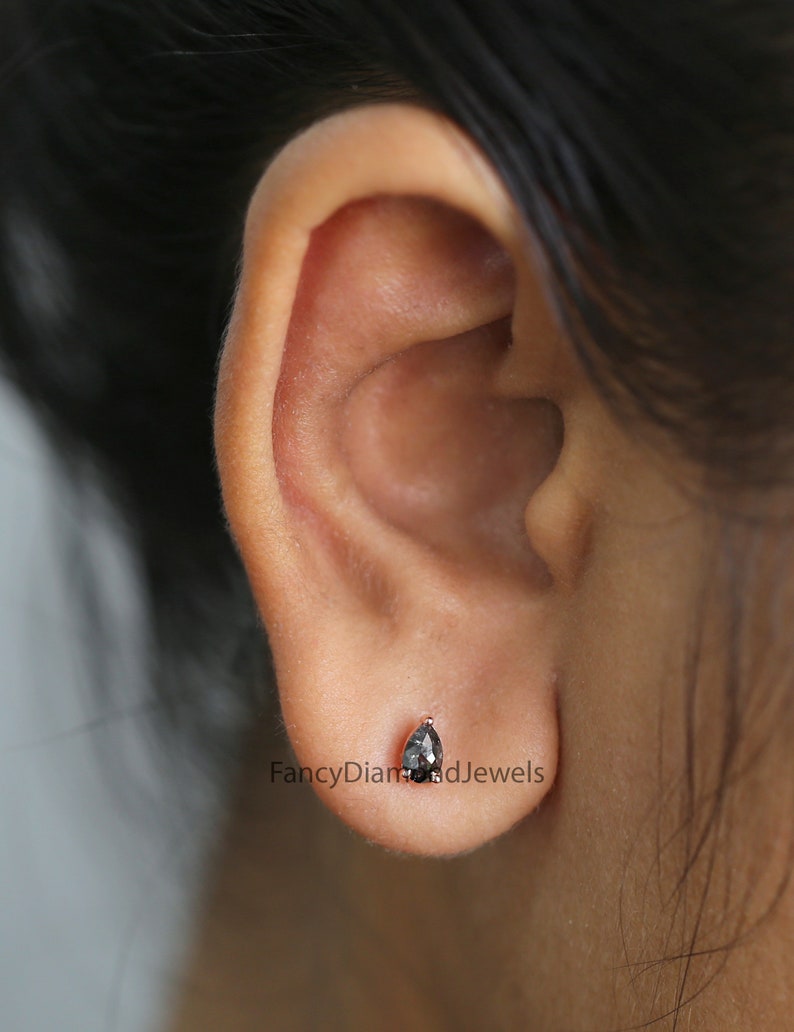 Puede incluir: Primer plano de una oreja con un peque&ntilde;o pendiente de diamante negro en forma de pera. El pendiente est&aacute; engastado en un engaste de plata u oro blanco.