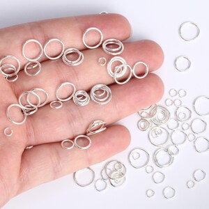 Op de afbeelding: Een verzameling zilverkleurige metalen buigringen in verschillende maten. De ringen zijn cirkelvormig, dun en hebben een kleine opening, verspreid over een oppervlak en rustend op een handpalm om de verschillende diameters te tonen.