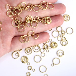 Op de afbeelding: Een verzameling goudkleurige metalen buigringen in verschillende maten. Deze ronde sieradenonderdelen hebben een eenvoudig open ontwerp, waardoor ze kunnen worden gebruikt als verbindingsstukken voor het maken van kettingen, armbanden en andere sieraden.