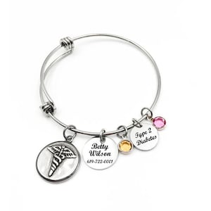 May include: A silver-tone expandable medical alert bangle bracelet featuring four charms: a circular caduceus emblem, a disc engraved with Betty Wilson and 619-722-0001, a yellow crystal charm, and a disc engraved with Type 2 Diabetes.