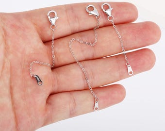 Può includere: Tre catenelle di estensione per gioielli color argento, ciascuna caratterizzata da un design a maglie sottili. Ogni catena è dotata di una chiusura a moschettone lucida a un'estremità e di una piccola targhetta metallica piatta e rettangolare.